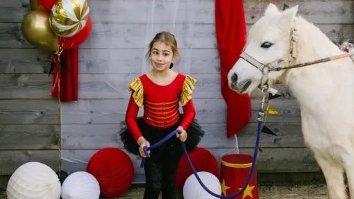 María, una niña de nueve años posando disfrazada de "domadora de circo" frente a un fondo de madera decorado con cortinas rojas y globos. A la izquierda de la fotografía su caballo blanco Txuri