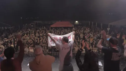 Vista de atrás de un escenario, sobre él aparecen personas sujetando una bandera en la que pone AEFAT y de fondo mucho público.