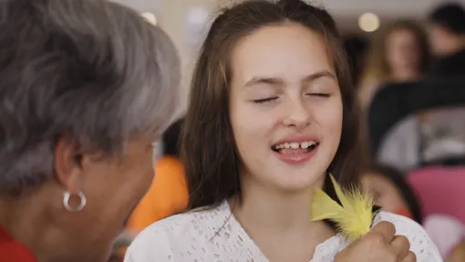 Fotografía en primer plano de una niña con síndrome de Rett con los ojos cerrados mientras una señora le hace cosquillas con una pluma.