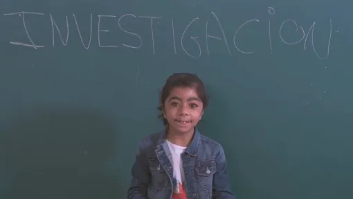Fotografía de una niña delante de una pizarra con la palabra "investigación" escrita en ella