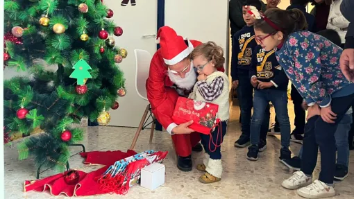 Fotografía: en el marco izquierdo, lateral de un árbol de navidad decorado con adornos. Centro de la imagen: Papá Noel entregando una bolsa navideña a una niña pequeña mientras, a su derecha, otros niños miran y esperan su turno.
