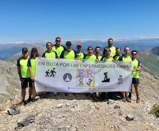 Personas con camiseta amarilla fosforito sujetando un cartel promocional de la asociación en ruta por las enfermedades raras en una montaña